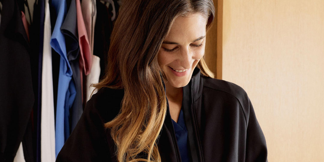 Image of woman putting on a scrub jacket by her closet. Her closet has a collection of scrubs.