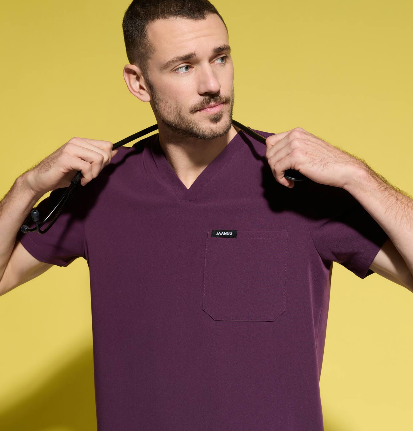 A man wearing JAANUU scrubs in Deep Eggplant color while holding a stethoscope around his neck.