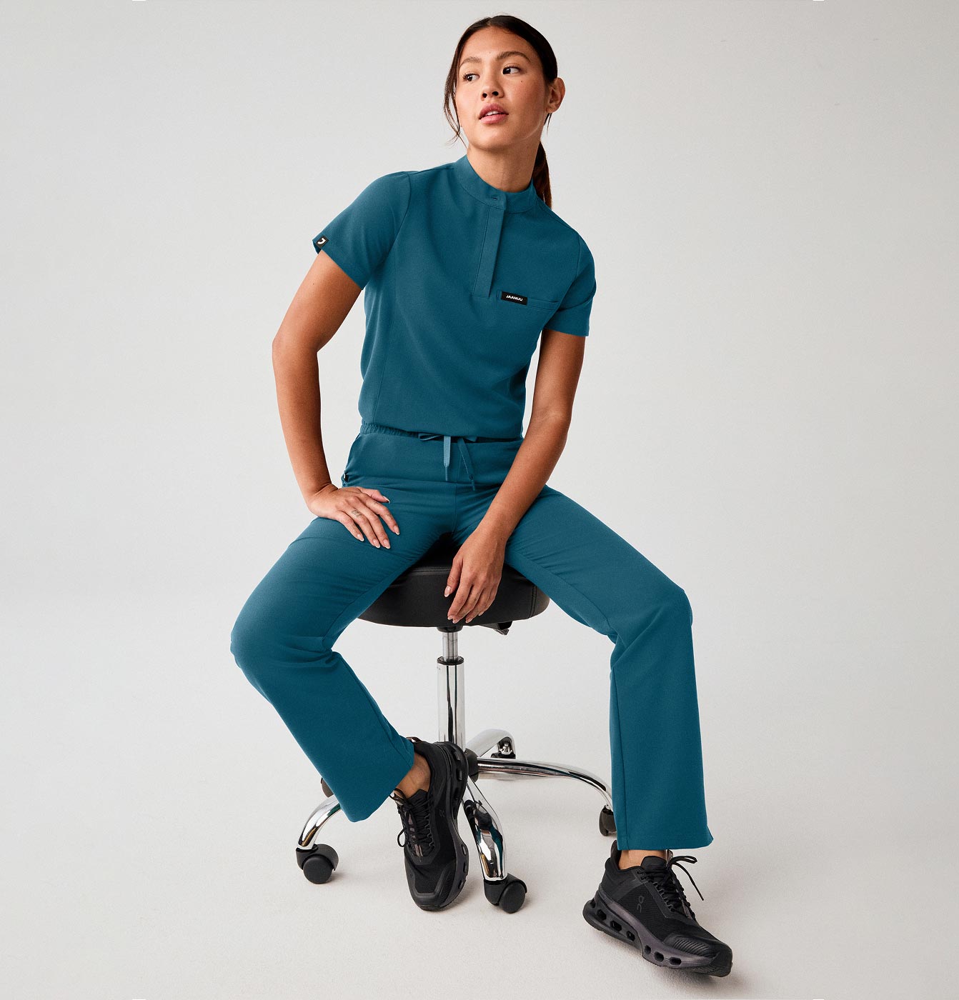 A woman wearing JAANUU scrubs sitting on a stool.