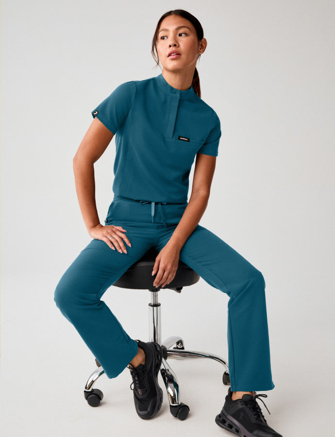 A woman wearing JAANUU scrubs sitting on a stool.
