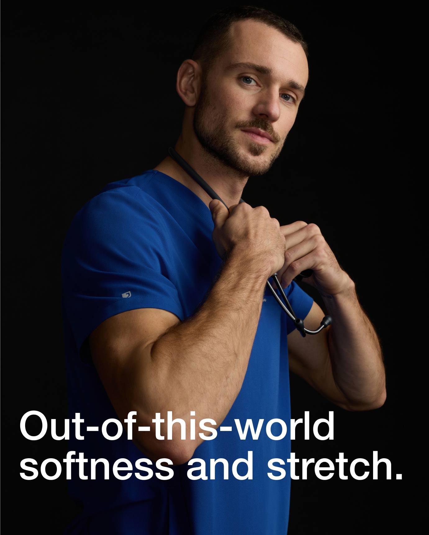 Image of a man wearing JAANUU Galaxy color scrubs with a stethoscope around his neck. "Image has text, "Out-of-this-world softness and stretch."