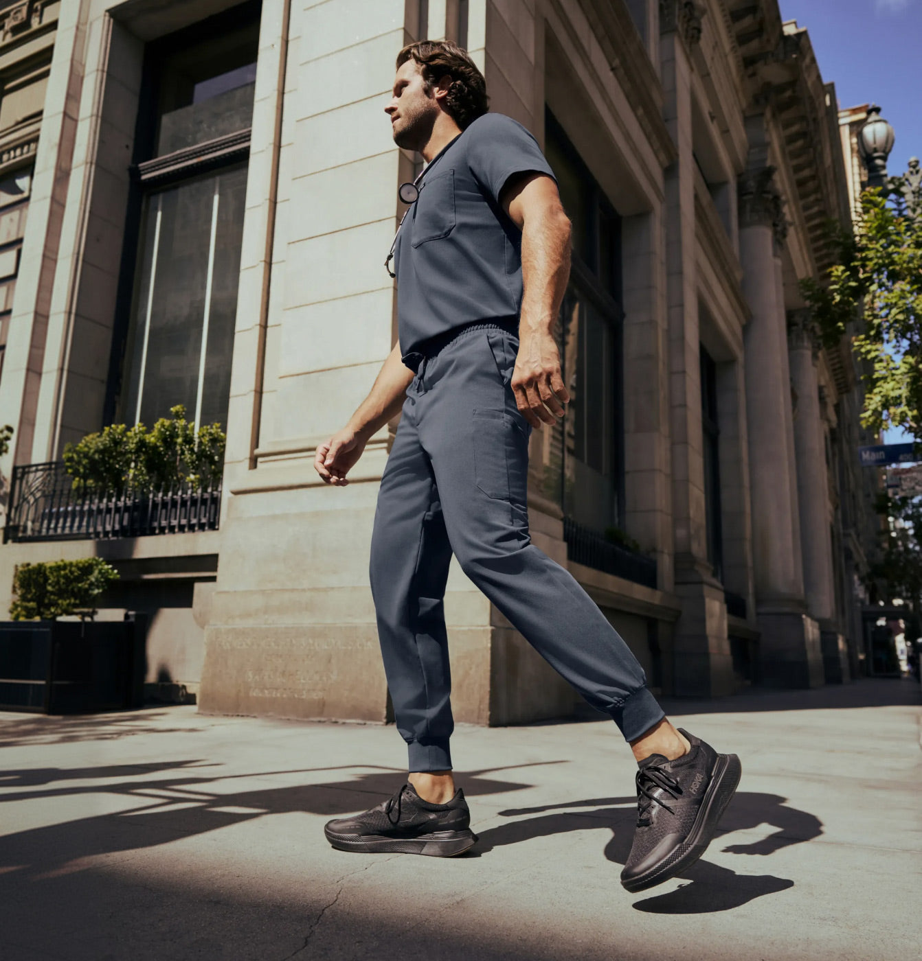 Man wearing JAANUU scrubs in Carbon Gray walking on a city sidewalk next to a building.