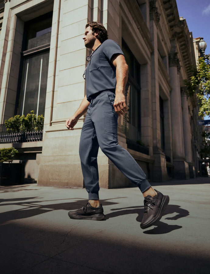 Man wearing JAANUU scrubs in Carbon Gray walking on a city sidewalk next to a building.
