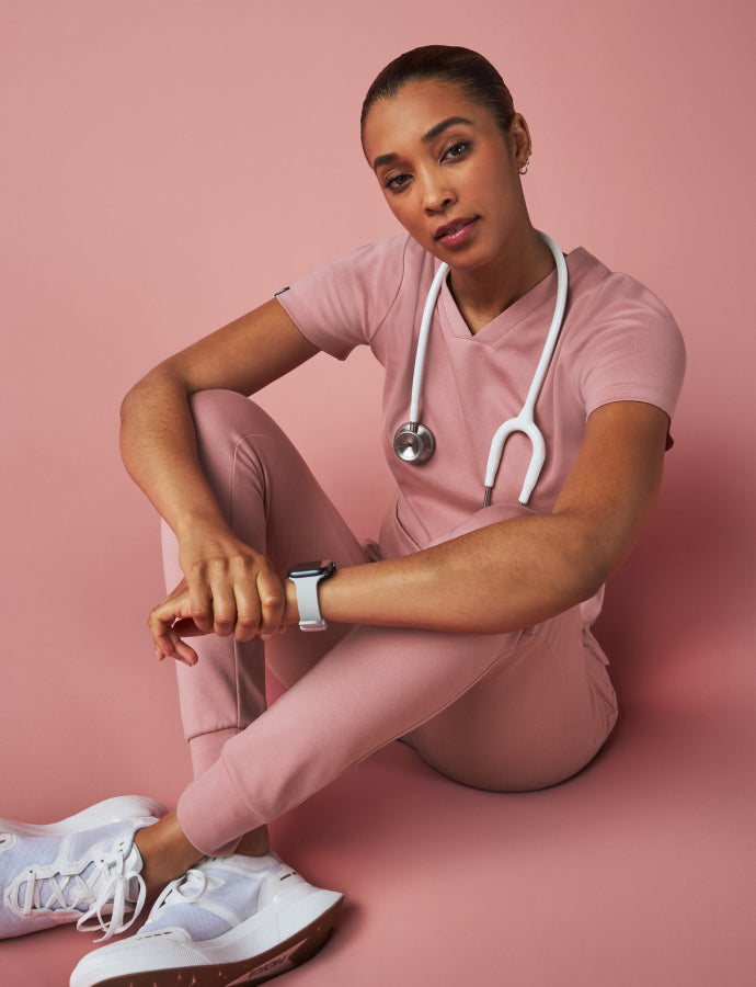 A women sitting down wearing Mauve scrubs with a stethoscope on a pink background