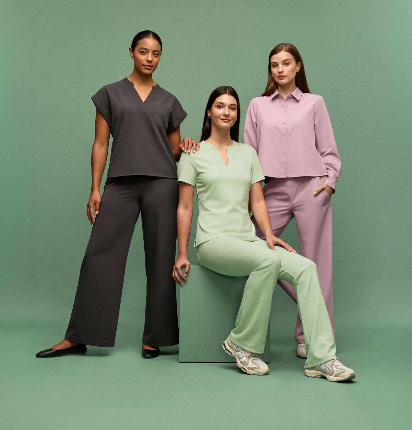 Three women wearing JAANUU scrub top and scrub pant in the same color: Carbon Gray, Mint, and Blossom on a green background