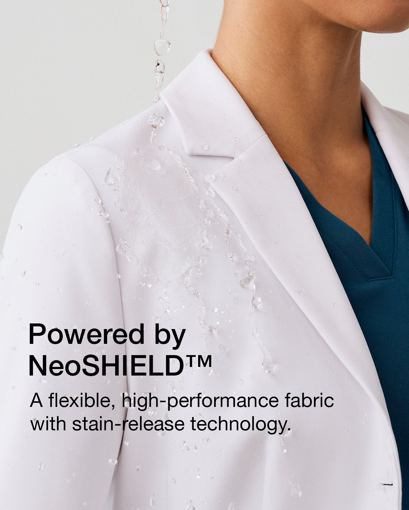 Close-up of a woman wearing a white Jaanuu lab coat with water droplets, with text 'Powered by NeoSHIELD™' and 'A flexible, high-performance fabric with stain-release technology.'