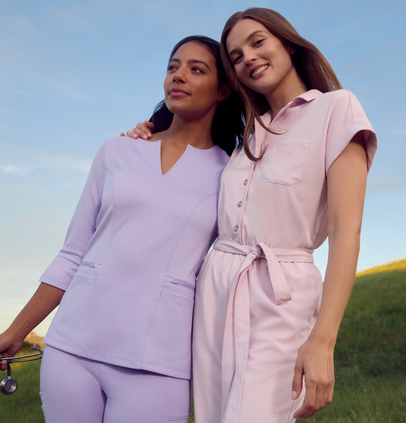 Two women standing together outdoors on a grassy hillside, one wearing Lavender Jaanuu scrubs holding a stethoscope and the other wearing Rose Quartz Jaanuu scrubs.