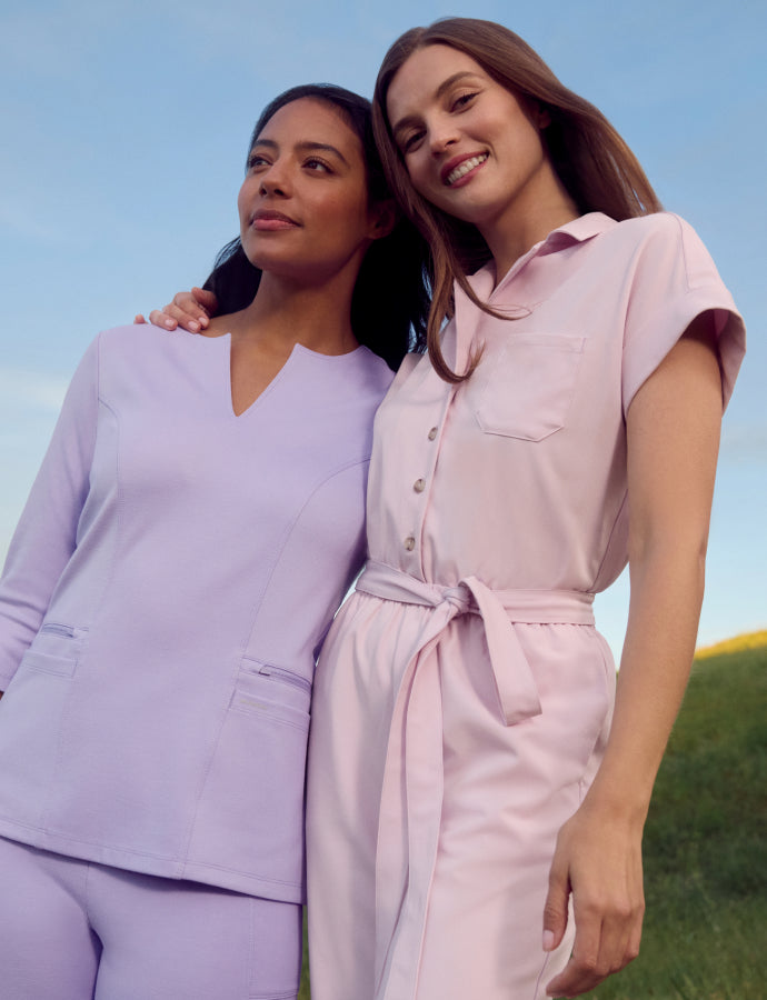 Two women standing together outdoors on a grassy hillside, one wearing Lavender Jaanuu scrubs holding a stethoscope and the other wearing Rose Quartz Jaanuu scrubs.
