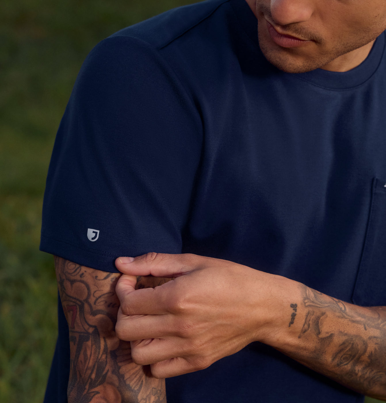 Close-up of a man wearing a navy Jaanuu UltraSOFT short-sleeve scrub top, with tattooed forearms on a green outdoor background