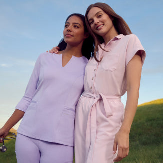 Two women standing together on a grassy hillside, one wearing a lavender Jaanuu scrub set holding a stethoscope and the other in a pink rose quartz Jaanuu button-front top and pants, against a clear blue sky - Women Nav Tile