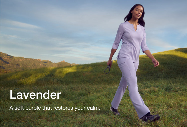 Woman walking through a grassy hillside wearing lavender Jaanuu scrubs and holding a stethoscope, with text 'Lavender' and 'A soft purple that restores your calm.'