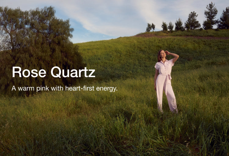 Woman standing on a grassy hillside wearing a rose quartz Jaanuu jumpsuit, with text 'Rose Quartz' and 'A warm pink with heart-first energy.'