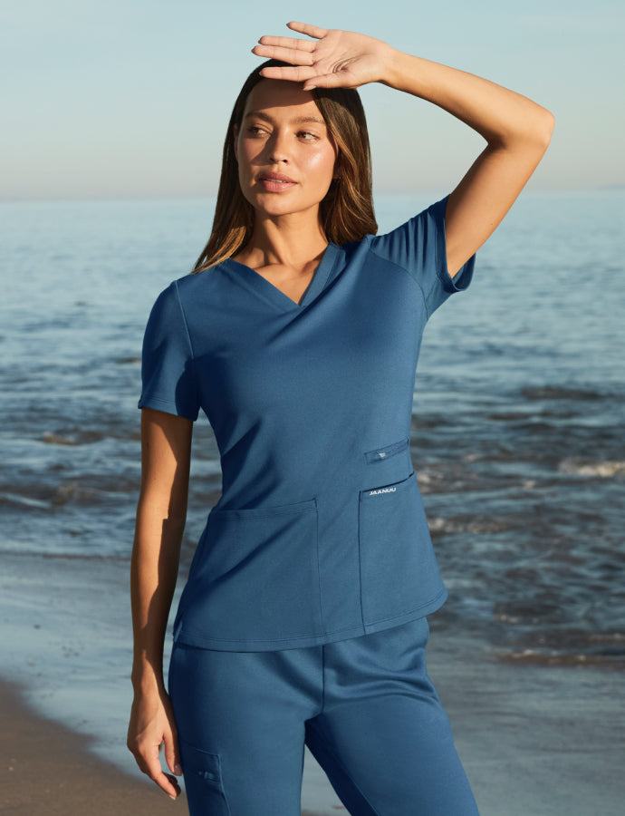 Woman wearing a blue Jaanuu scrub set standing on a beach with the ocean in the background, hand shielding her eyes