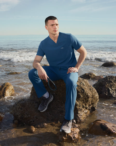 Man in blue Jaanuu scrubs sitting on a rock at the beach, holding a stethoscope - Men Nav Tile