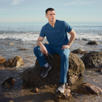 Man in blue Jaanuu scrubs sitting on a rock at the beach, holding a stethoscope - Men Nav Tile