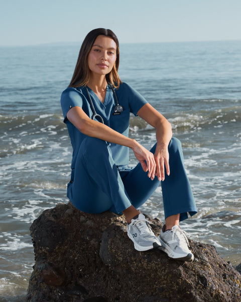 Woman in blue Jaanuu scrubs sitting on a rock at the beach, looking at the camera - Women Nav Tile