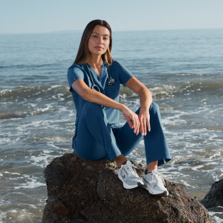 Woman in blue Jaanuu scrubs sitting on a rock at the beach, looking at the camera - Women Nav Tile