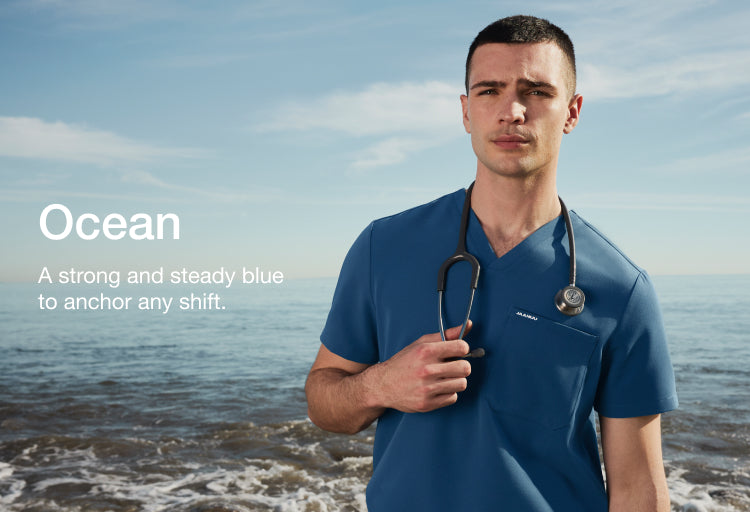 Man wearing a blue Jaanuu scrub top with stethoscope on a beach, with text 'Ocean' and 'A strong and steady blue to anchor any shift.'