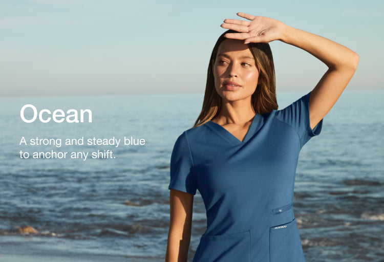 Woman wearing a blue Jaanuu scrub top on a beach, hand shielding her eyes, with text 'Ocean' and 'A strong and steady blue to anchor any shift.'