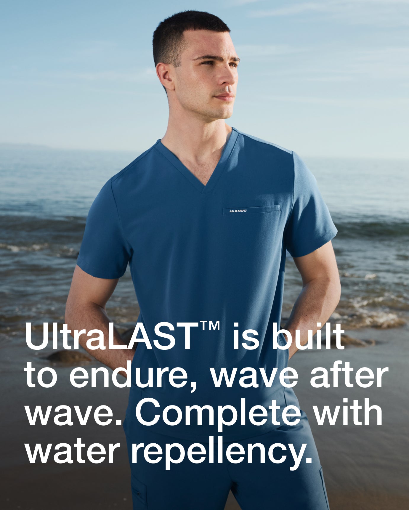 Man wearing a blue Jaanuu scrub set on a beach, with text 'UltraLAST is built to endure, wave after wave. Complete with water repellency.'