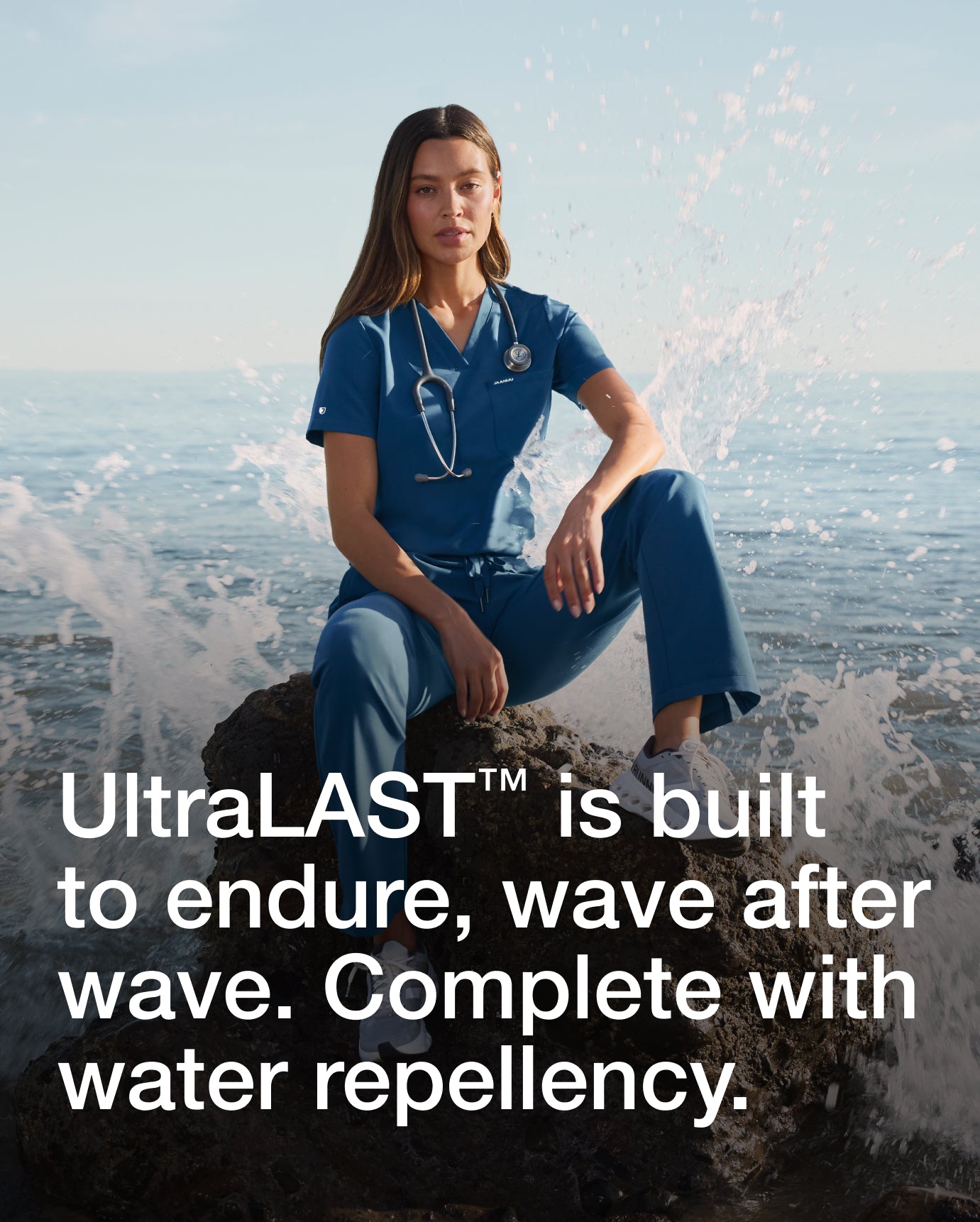 Woman wearing a blue Jaanuu scrub set sitting on a rock with ocean spray behind her, with text 'UltraLAST is built to endure, wave after wave. Complete with water repellency.'