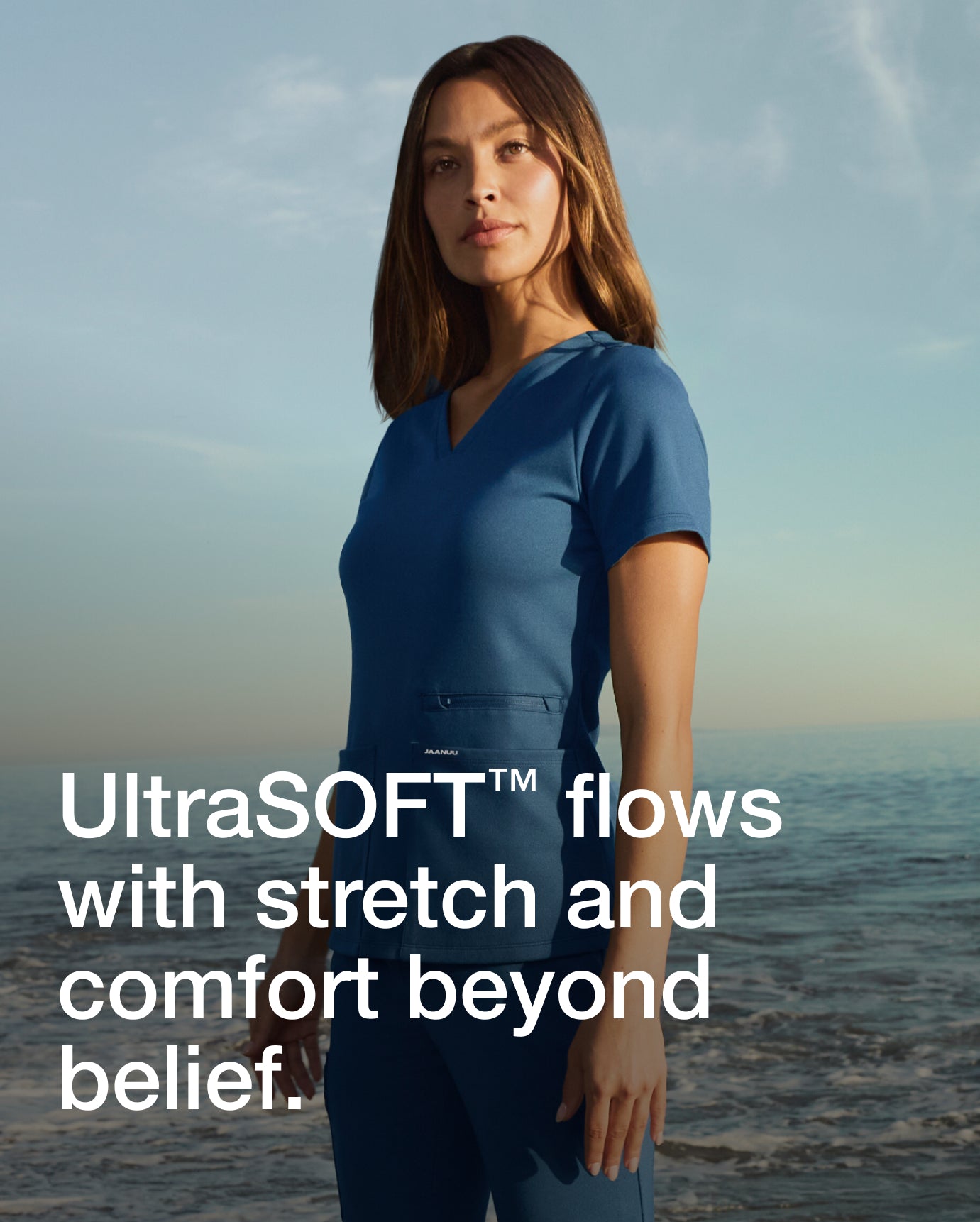 Woman wearing a blue Jaanuu scrub top on a beach, with text 'UltraSOFT flows with stretch and comfort beyond belief.'