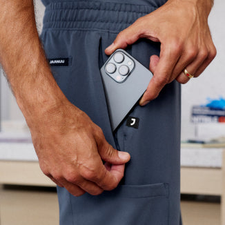 Man wearing carbon gray scrub pants with a JAANUU logo, inserting a phone to the pocket - Men Nav Tile