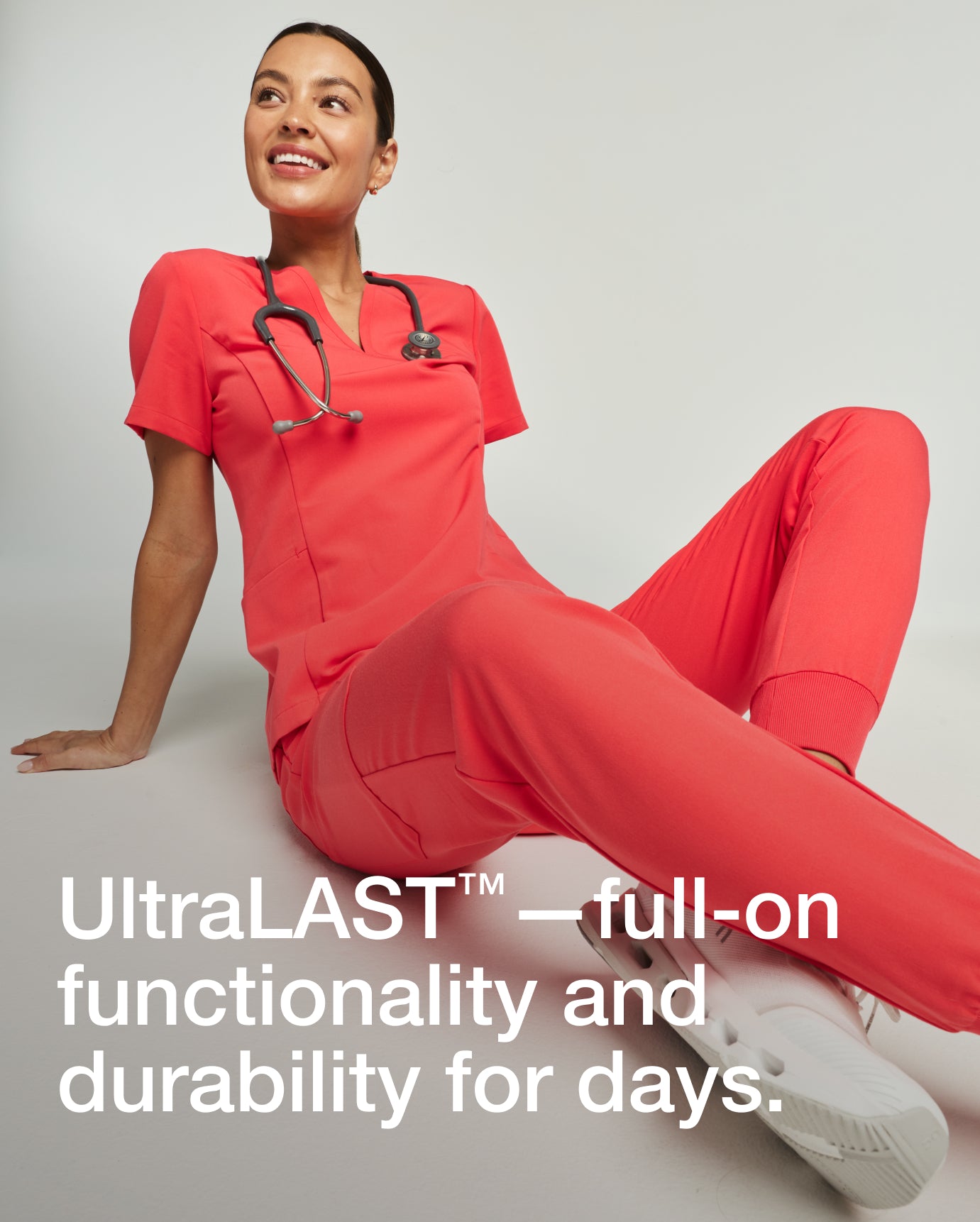 Woman wearing a coral Jaanuu scrub set with stethoscope, seated on the floor and smiling, with text 'UltraLAST™—full-on functionality and durability for days.'