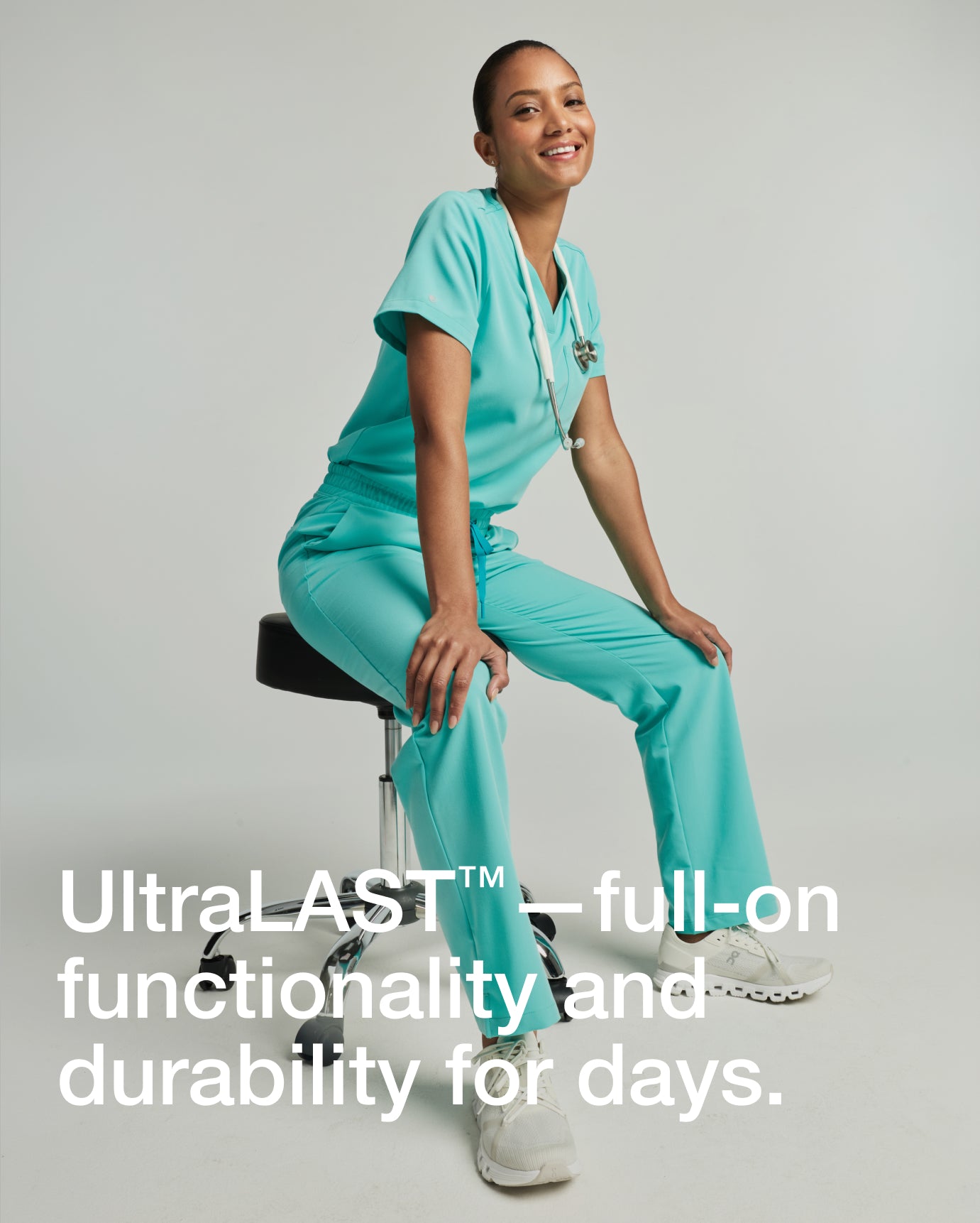 Woman wearing a surf teal Jaanuu scrub set seated on a stool with stethoscope, smiling, with text 'UltraLAST™—full-on functionality and durability for days.'