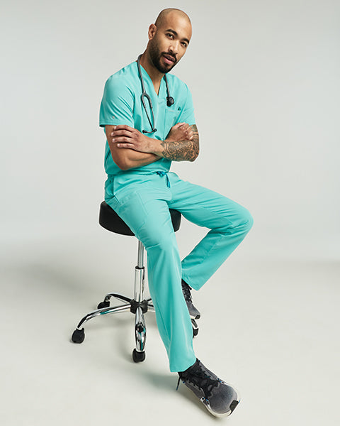 Man wearing a surf teal Jaanuu scrub top and woman wearing a coral Jaanuu scrub top standing together, with text '25% Off Sitewide' - Men Nav Tile