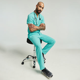 Man wearing a surf teal Jaanuu scrub top and woman wearing a coral Jaanuu scrub top standing together, with text '25% Off Sitewide' - Men Nav Tile