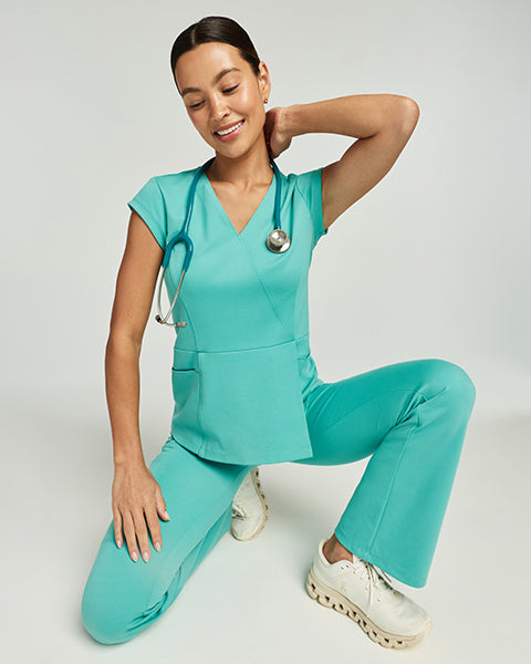 Man wearing a surf teal Jaanuu scrub top and woman wearing a coral Jaanuu scrub top standing together, with text '25% Off Sitewide' - Women Nav Tile