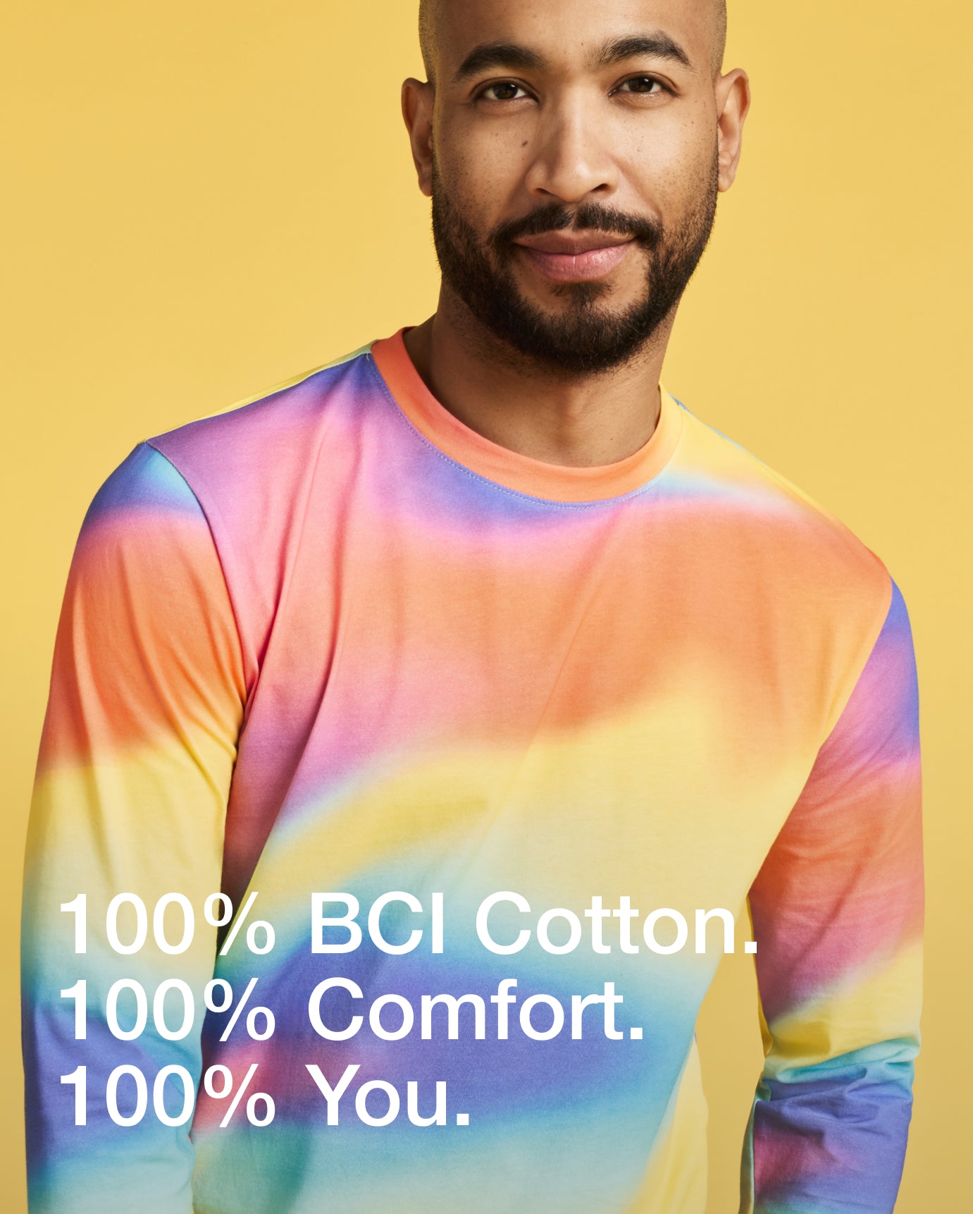 Man wearing a rainbow tie-dye Jaanuu Pride underscrub on a yellow background, with text '100% BCI Cotton. 100% Comfort. 100% You.'