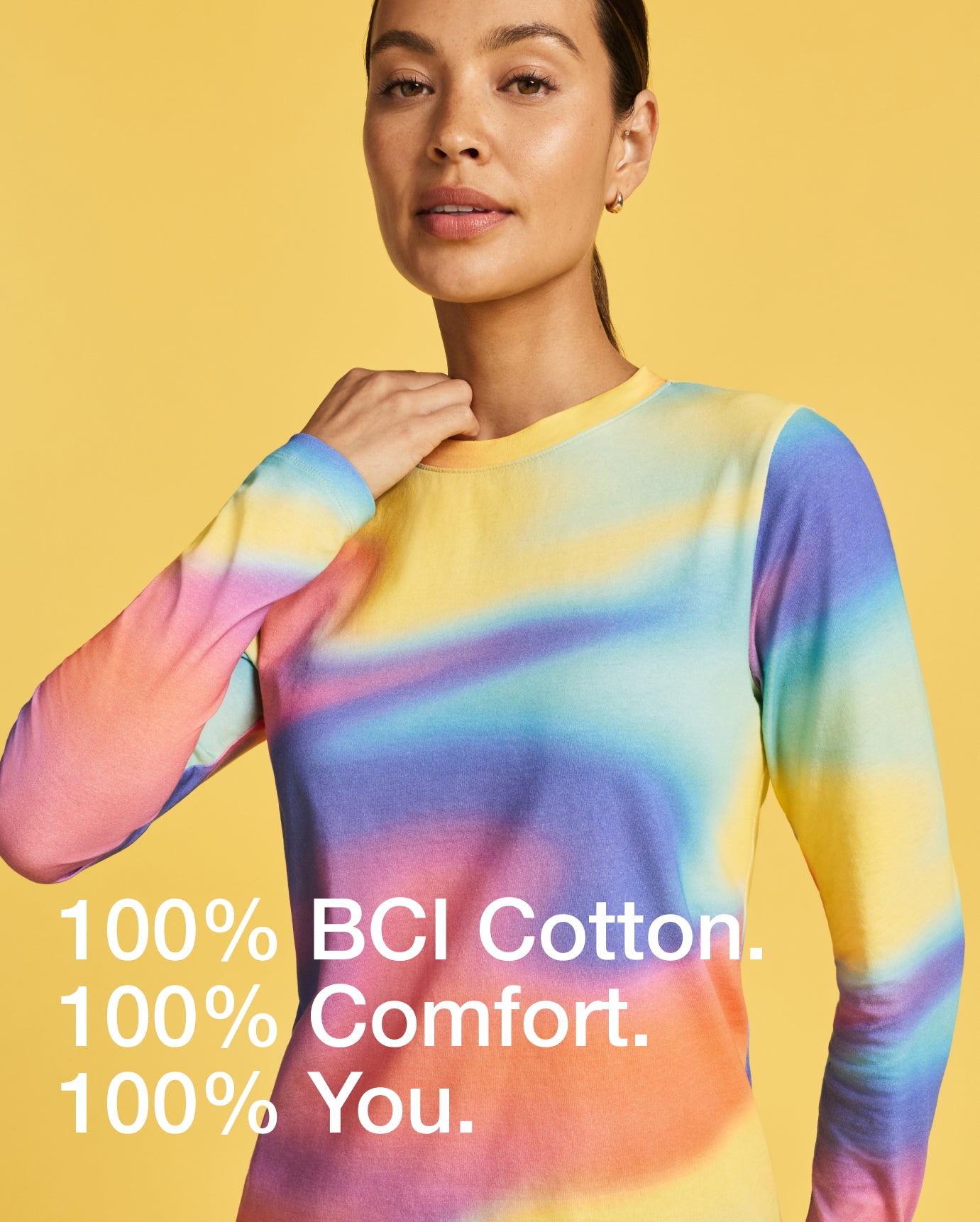 Woman wearing a rainbow tie-dye Jaanuu Pride underscrub on a yellow background, with text '100% BCI Cotton. 100% Comfort. 100% You.'