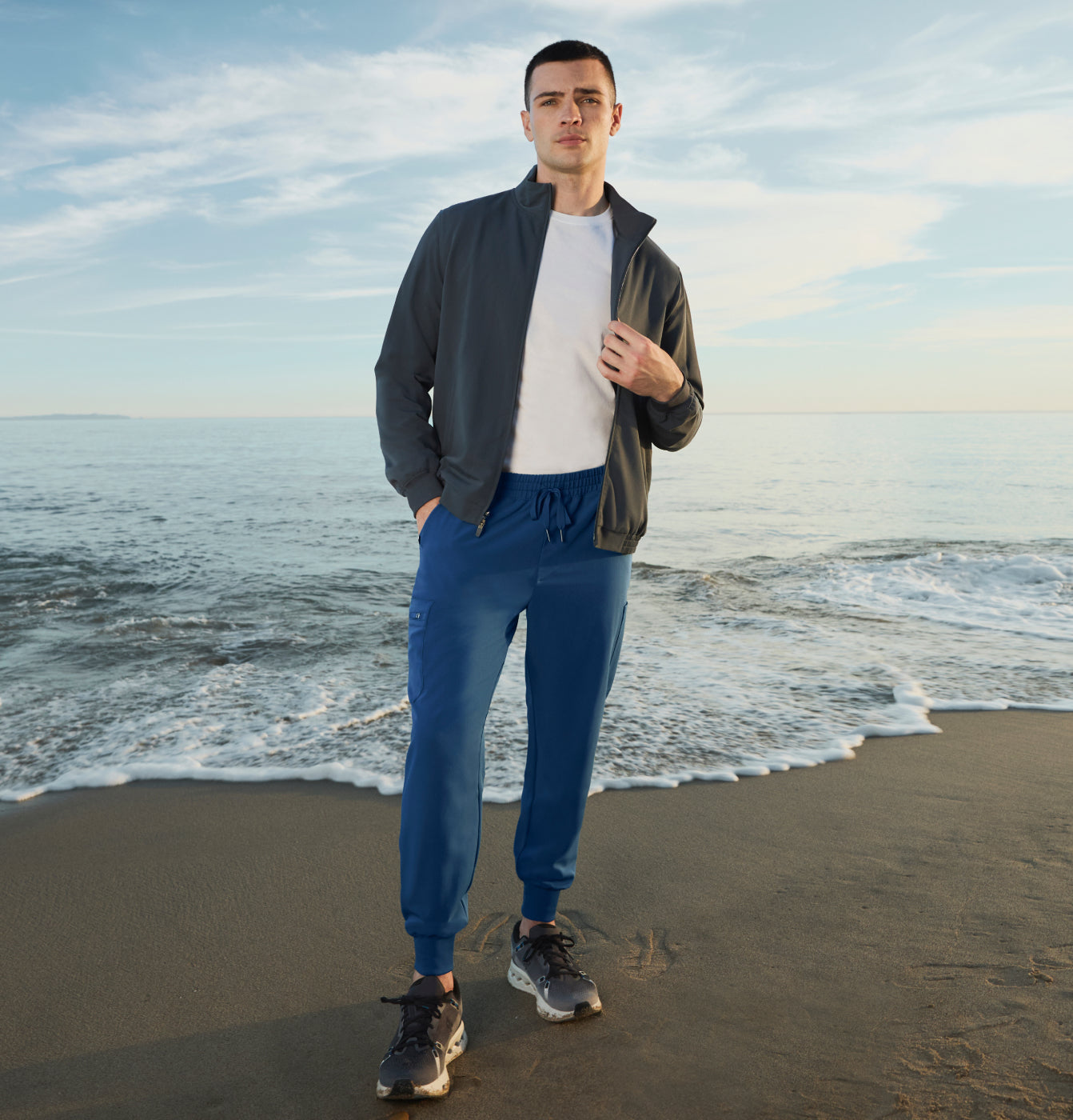 Man standing on the beach wearing a charcoal jacket over a white tee with blue jogger scrub pants
