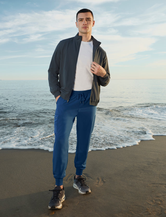 Man standing on the beach wearing a charcoal jacket over a white tee with blue jogger scrub pants
