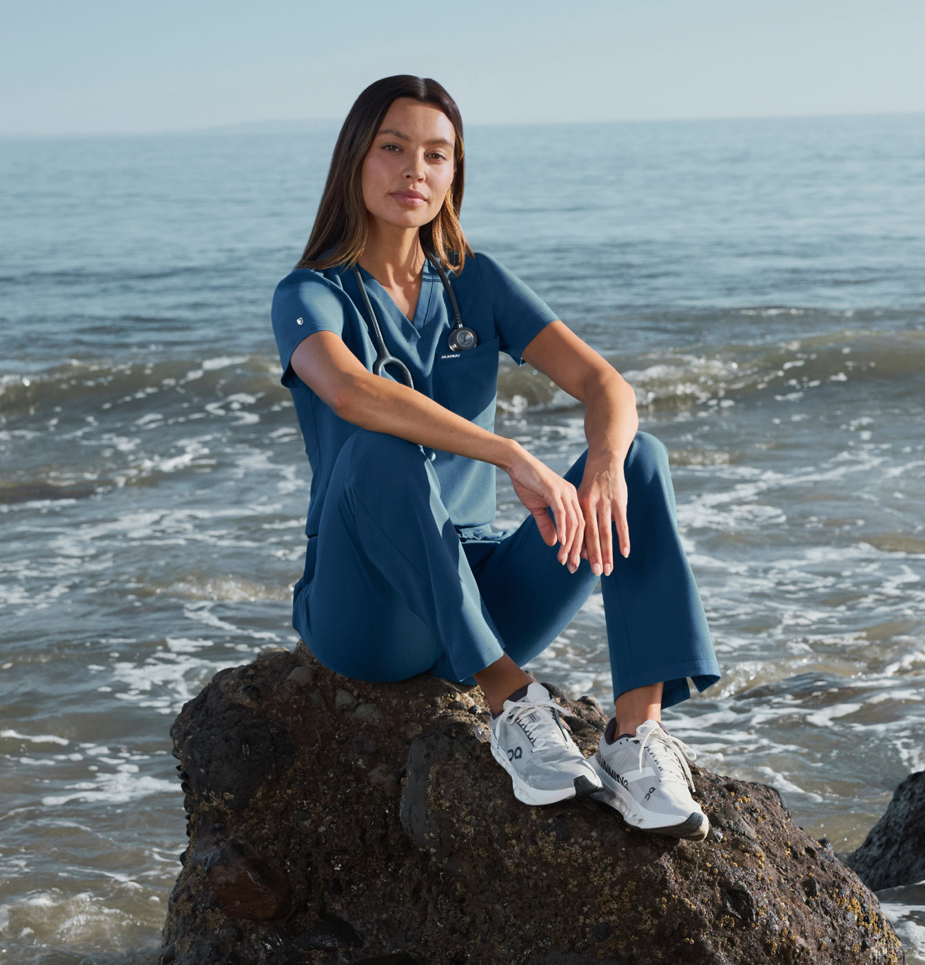 Woman in ocean blue scrubs sitting on a beach rock with waves rolling in behind her