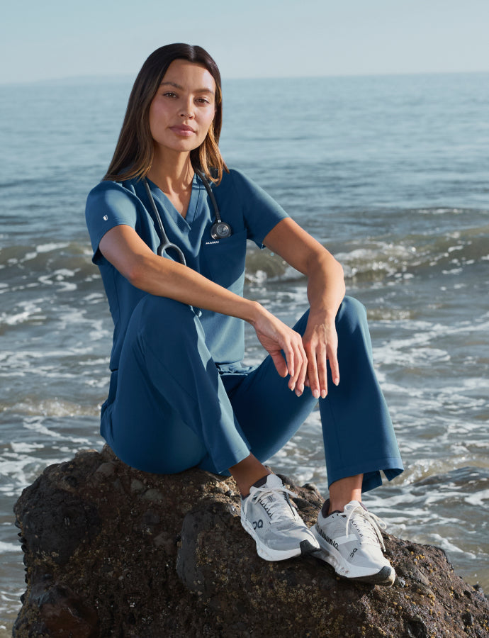 Woman in ocean blue scrubs sitting on a beach rock with waves rolling in behind her