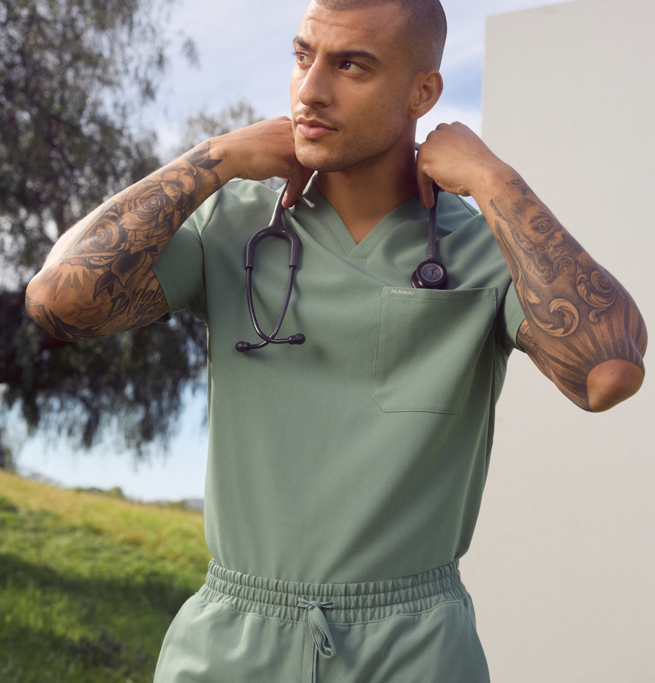 Man in sage green scrubs with tattooed forearms holding a stethoscope behind his neck, standing outdoors by a tree