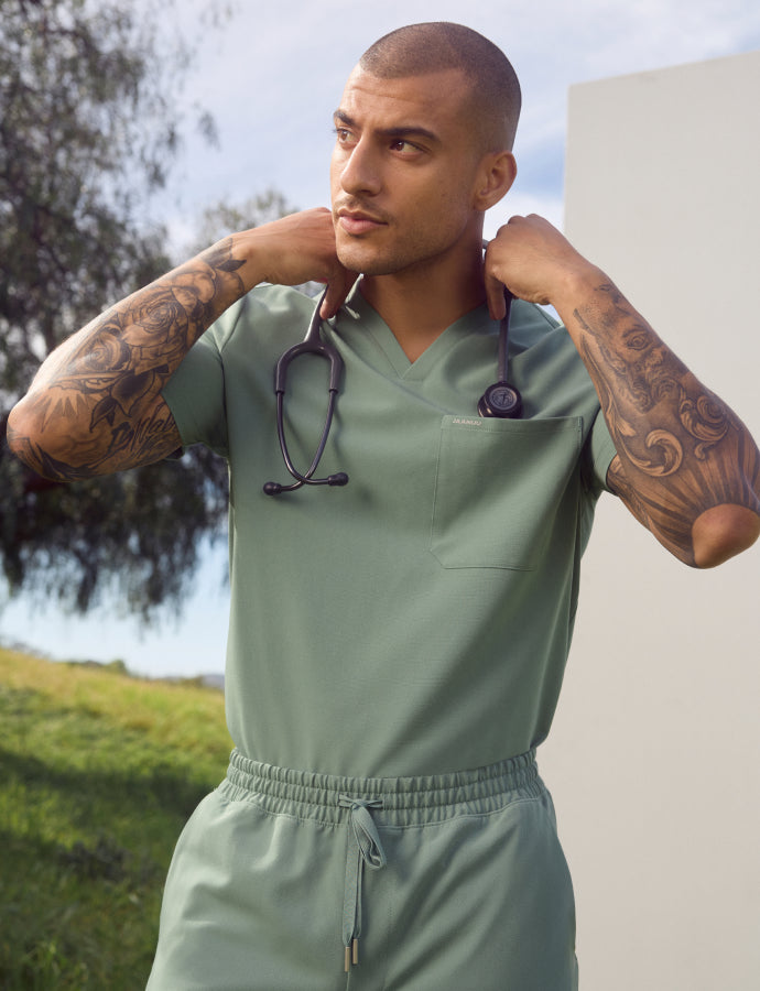Man in sage green scrubs with tattooed forearms holding a stethoscope behind his neck, standing outdoors by a tree