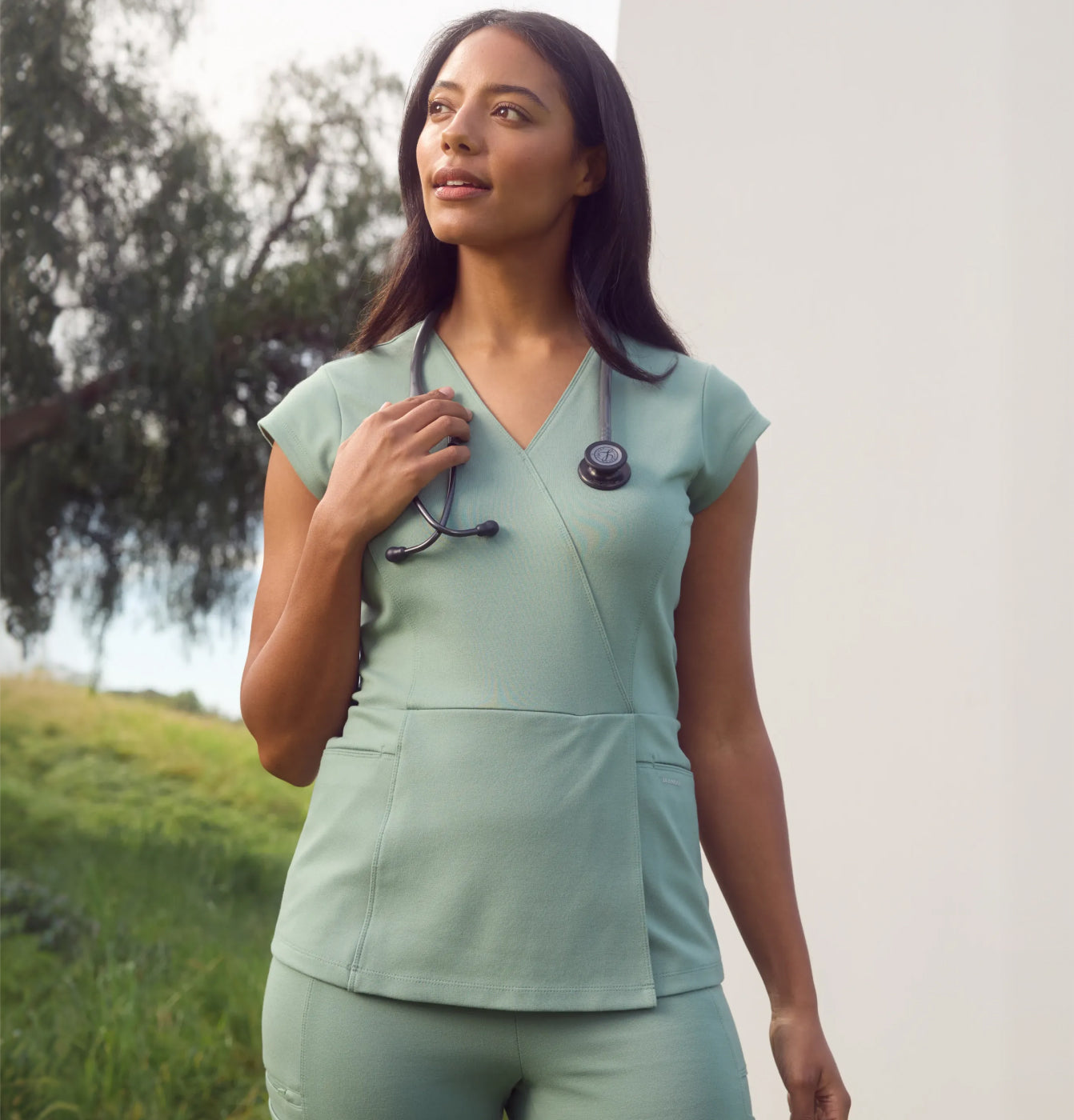 Woman in sage green scrubs with a stethoscope around her neck, standing in front of a tree and a grassy hillside