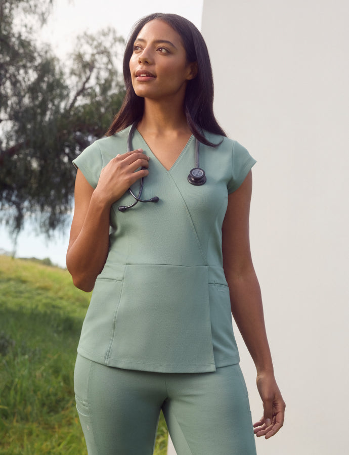 Woman in sage green scrubs with a stethoscope around her neck, standing in front of a tree and a grassy hillside