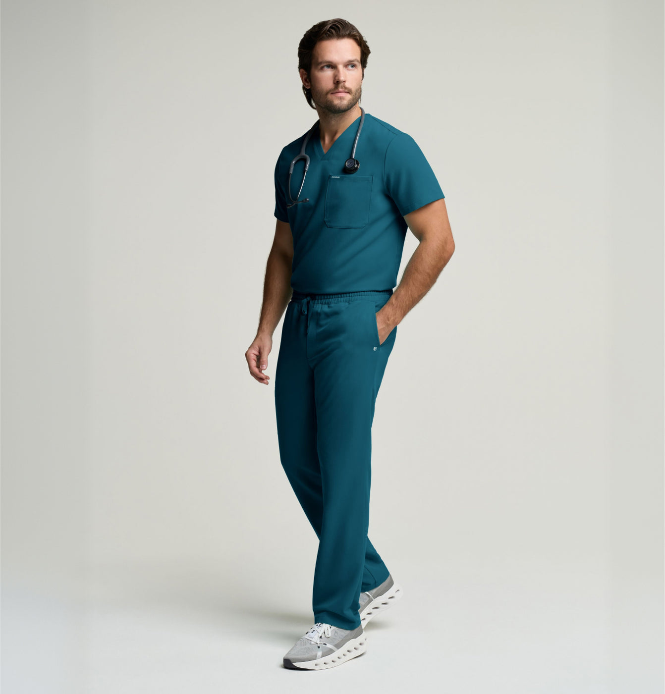 Man in dark teal scrubs with a stethoscope around his neck, standing in a studio