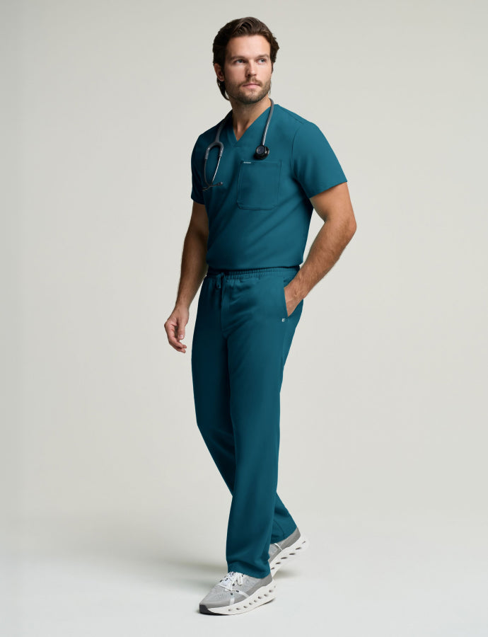 Man in dark teal scrubs with a stethoscope around his neck, standing in a studio