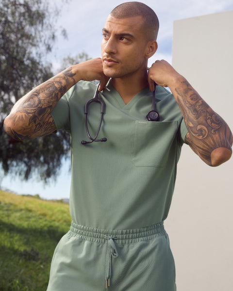 A man with a shaved head and tattooed arms wearing a sage green Jaanuu scrub top with a stethoscope, outdoors against green hills. - Men Nav Tile