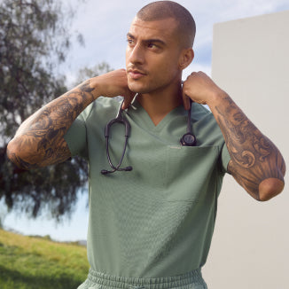 A man with a shaved head and tattooed arms wearing a sage green Jaanuu scrub top with a stethoscope, outdoors against green hills. - Men Nav Tile