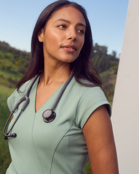 A woman with dark hair wearing a sage green Jaanuu scrub top with a stethoscope around her neck, outdoors against green hills. - Women Nav Tile