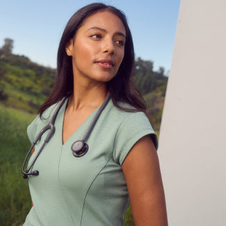 A woman with dark hair wearing a sage green Jaanuu scrub top with a stethoscope around her neck, outdoors against green hills. - Women Nav Tile