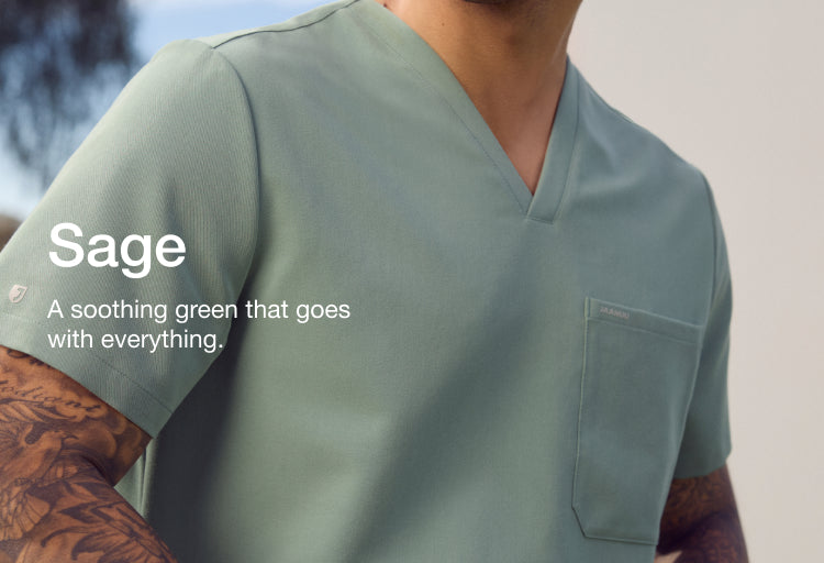 A close-up of a man in a sage green Jaanuu V-neck scrub top with a tattooed forearm, against a blurred green hillside. Text on the image reads: "Sage. A soothing green that goes with everything."