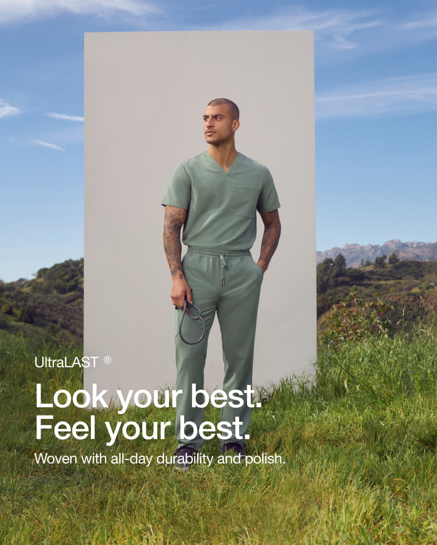 A man in a sage green Jaanuu UltraLAST scrub set holds a stethoscope on a grassy hillside. Text on the image reads: "Look your best. Feel your best. Woven with all-day durability and polish." with an UltraLAST fabric label.